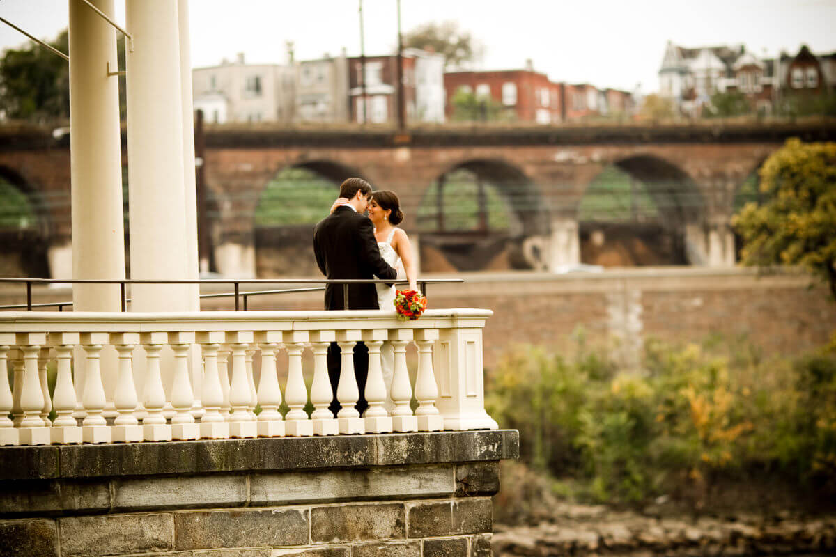 Elegant Wedding Couple Pose For A Portrait With The Buildings And Bridge In The Background